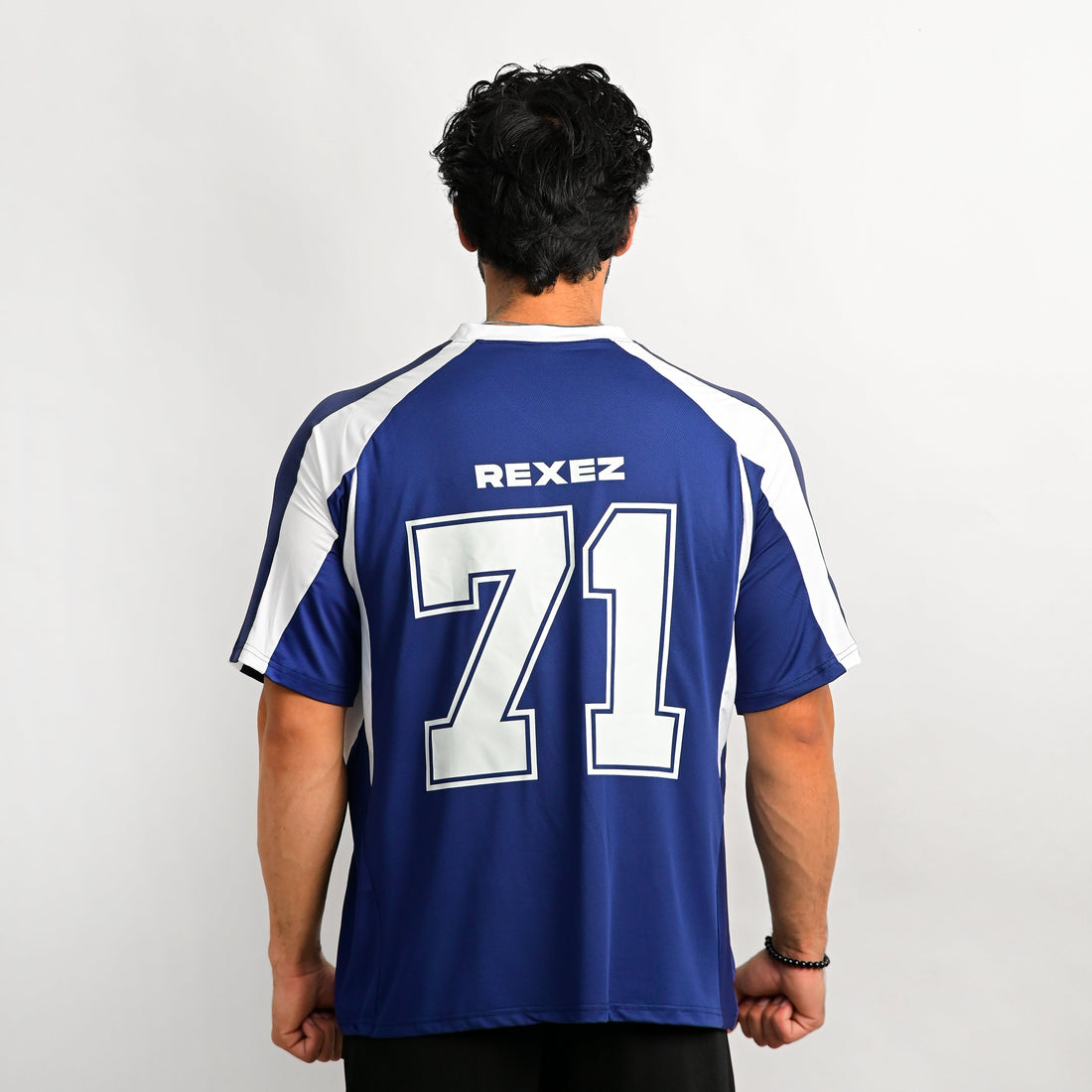 A man viewed from behind wearing a royal blue athletic jersey with white shoulder panels and the name REXEZ and number 71 printed on the back, against a plain light gray studio background.